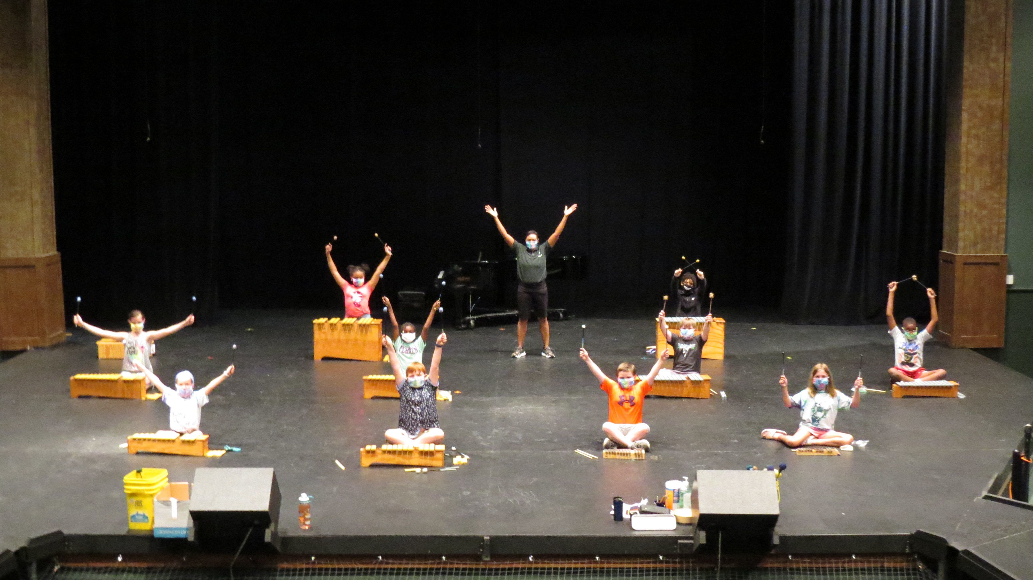 A socially distanced group of elementary aged students and their instructors pose on a large stage with their orff instruments