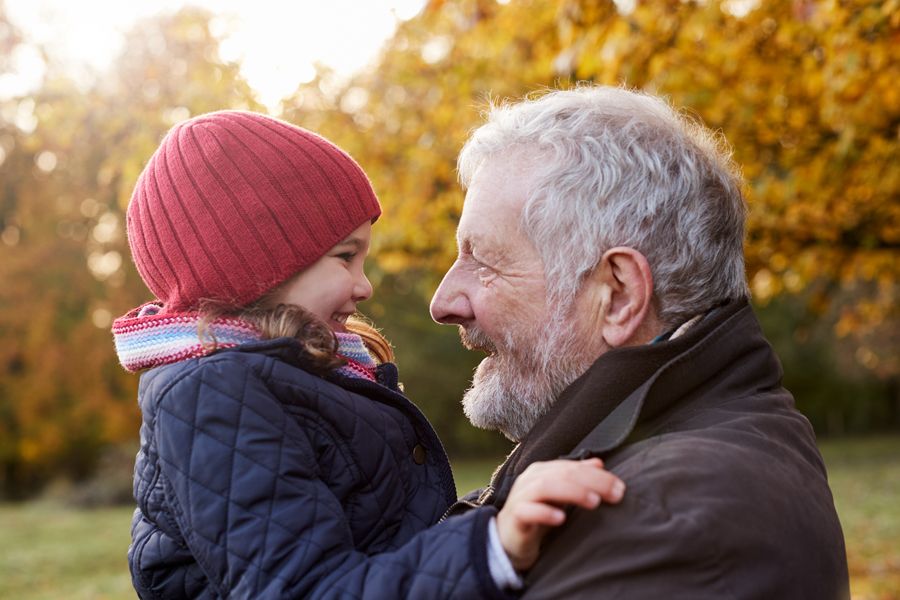 Grandfather holding 4-year-old granddaughter outside during the fall, both are smiling