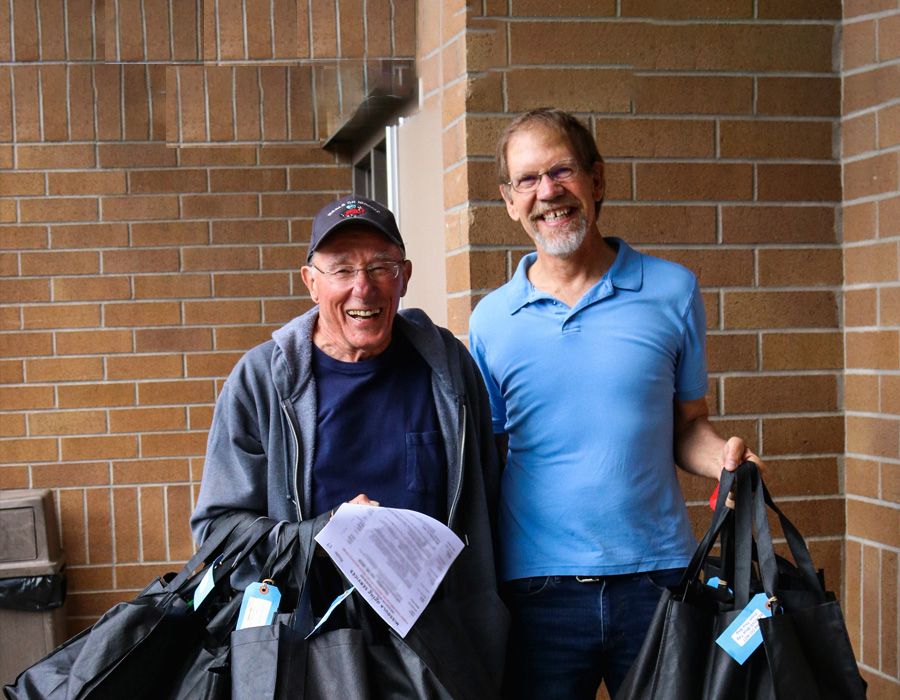 two older men smiling while holding bags to deliver meals for meals on wheels
