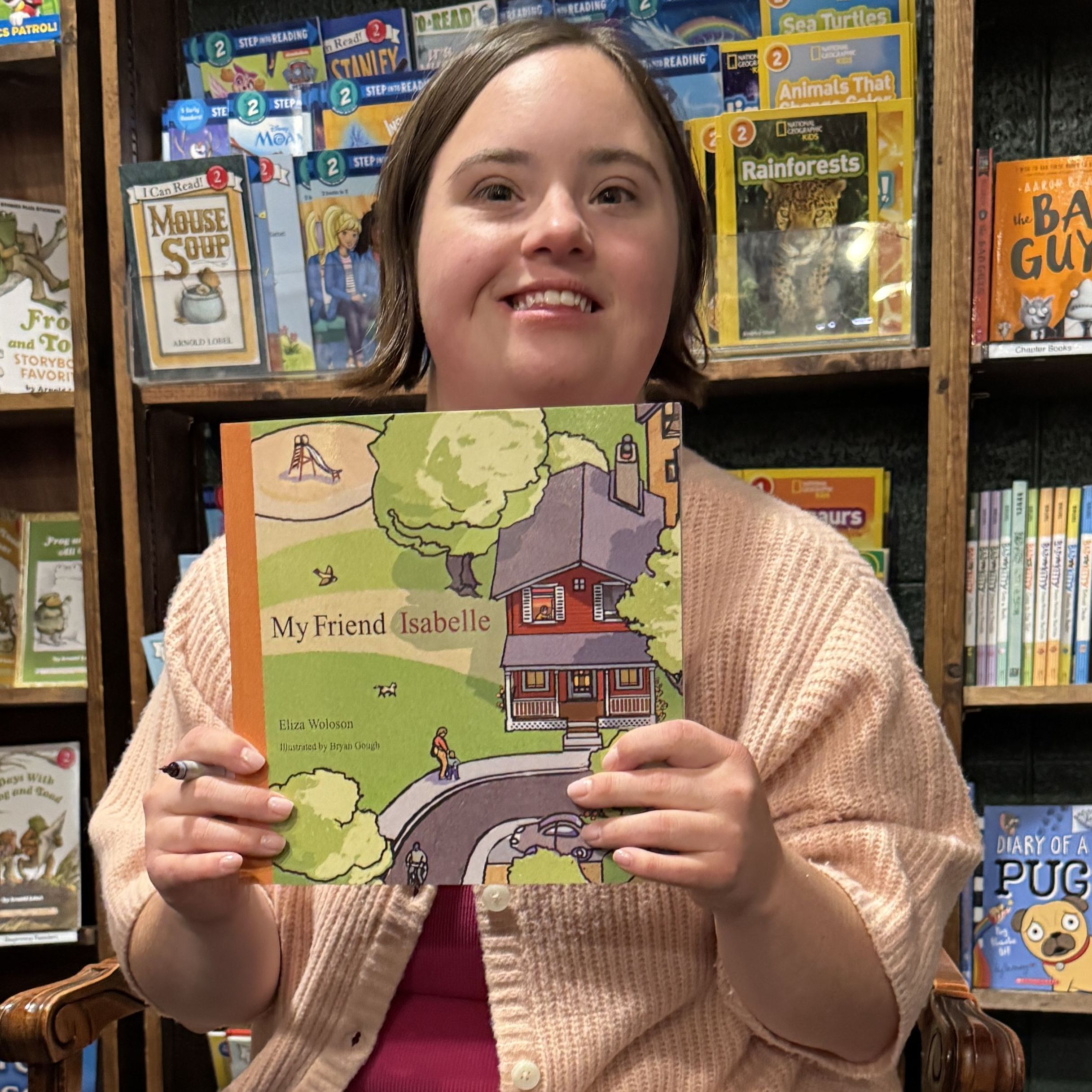 Isabelle is a white woman with Down syndrome. She sits in a chair wearing a pink cardigan. She smiles and holds up the picture book "My Friend Isabelle."