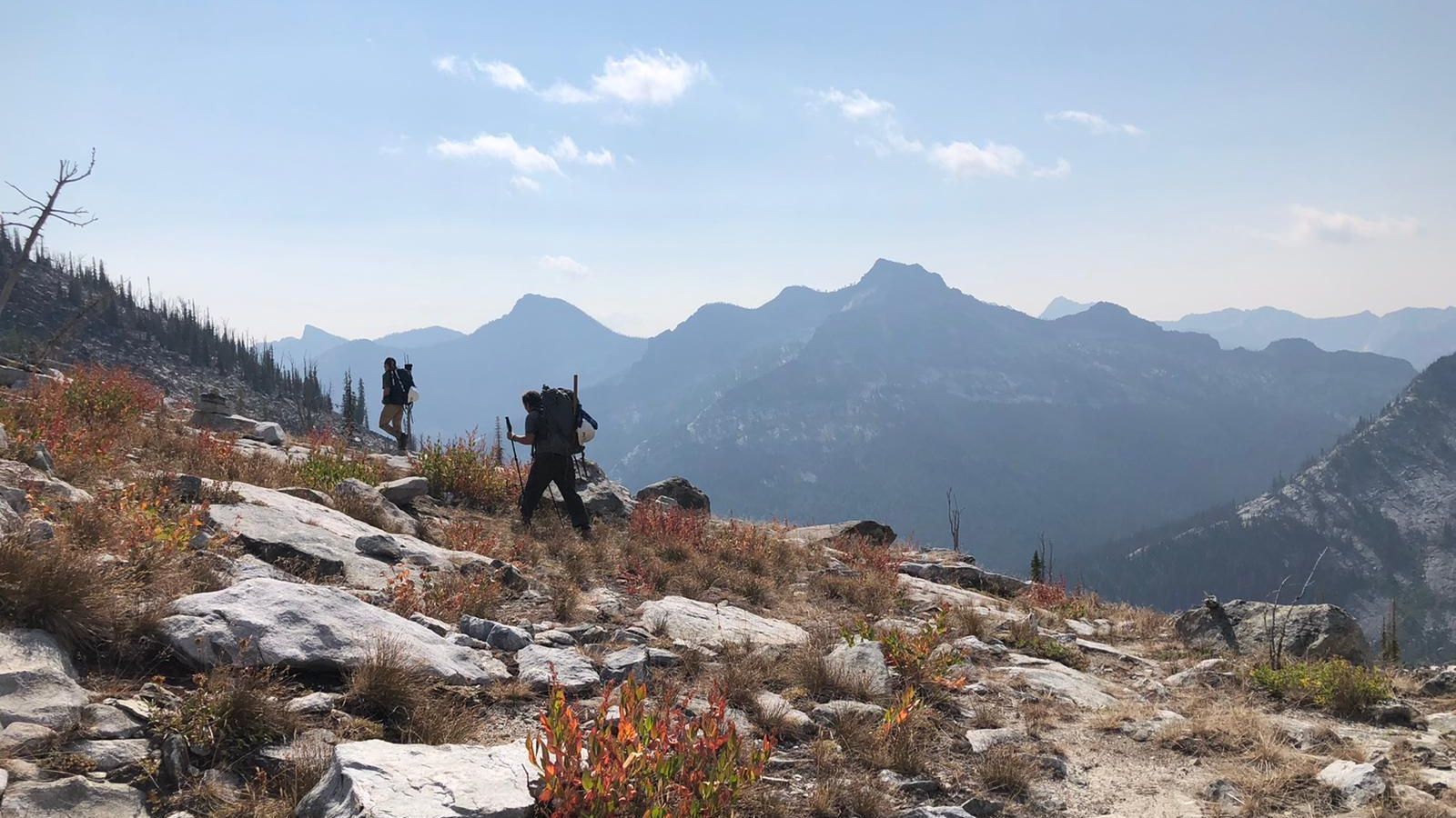 [Image Description: Two MCC members are walking away on a rocky trail, carrying their packs, surrounded by burnt orange bushes. Through the haze in the background, there are a multitude of mountains, overlapping one another.]