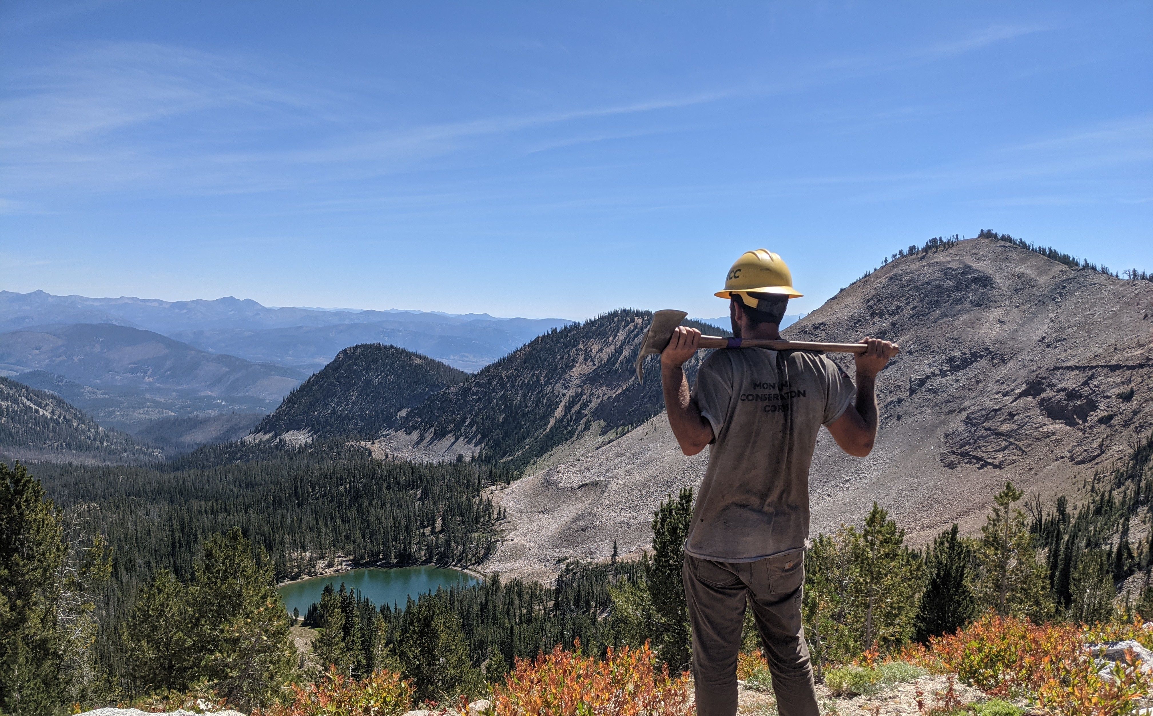 [Image Description: An MCC member, wearing their hard hat and dirt-covered uniform, is looking over a lake, surrounded by mountains of scree rock. They are holding an axe over their shoulders, taking in the beauty of the landscape.]