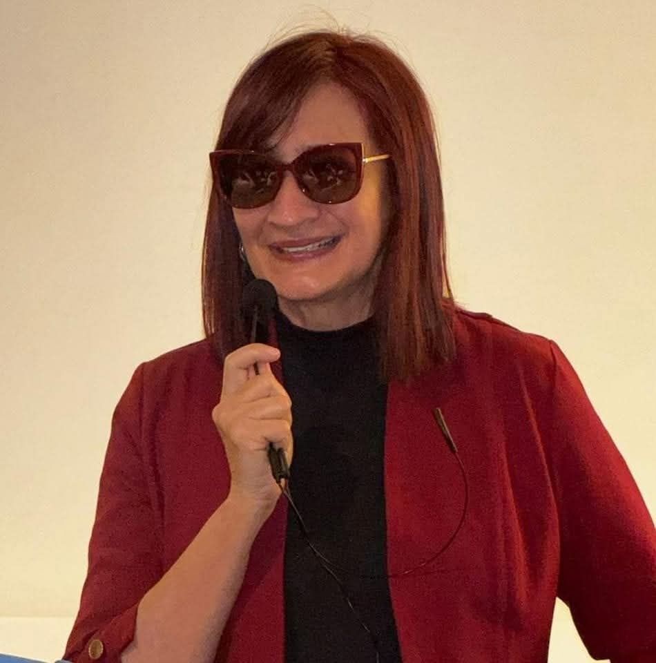 Woman with red hair and sunglasses speaks into a handheld microphone.
