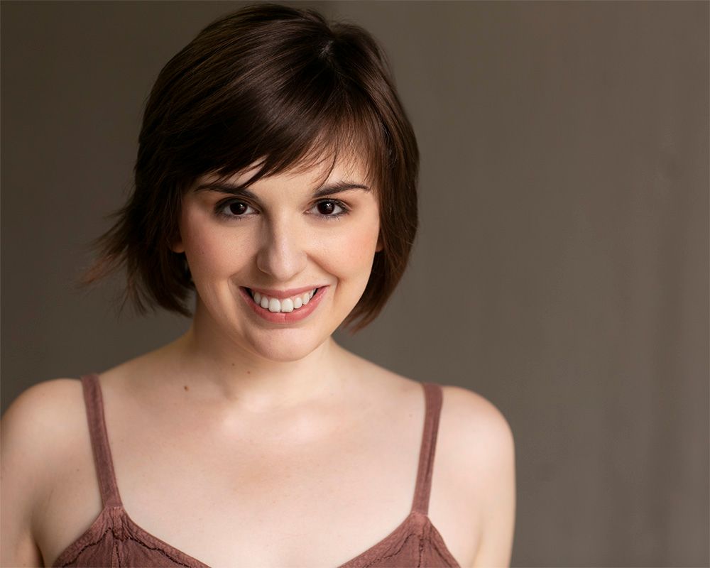 Bree Klauser, a women who is in her 20's-30's. She is wearing a brown blouse, looking straight at the camera smiling with a dark back drop.