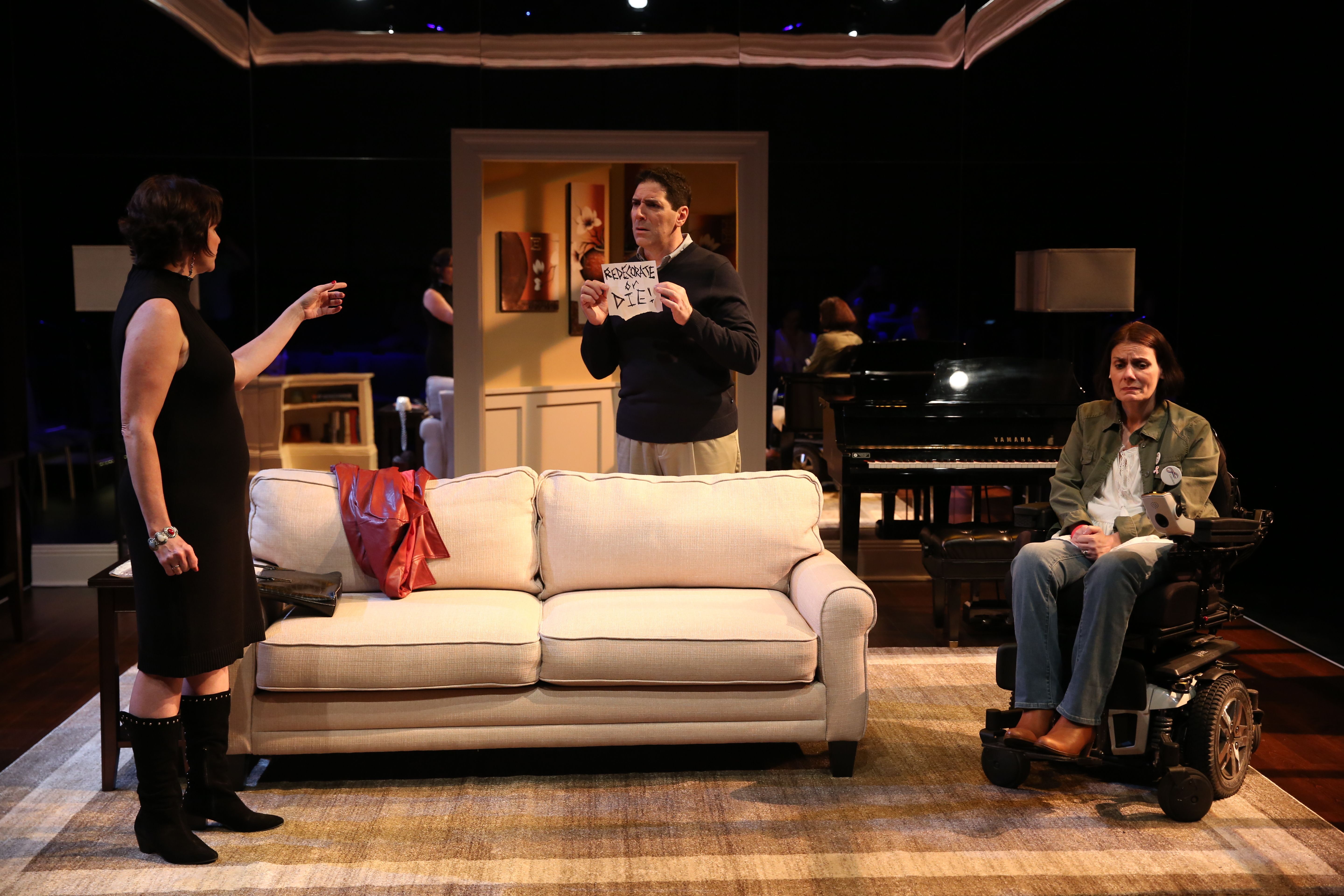 A picture of three actors who’re both sitting and standing, and they’re performing a scene from The Fourth Wall. The actor on the left is pointing her finger to the middle actor who is holding up a sign. The actor on the right is looking sad.