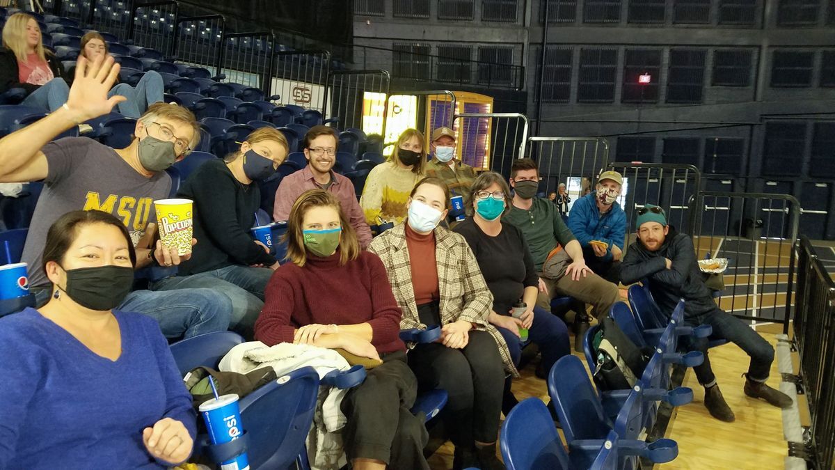 [Image Description: Bozeman State Staff sit on the bleachers in an arena, watching a basketball game. They are wearing facemasks.]