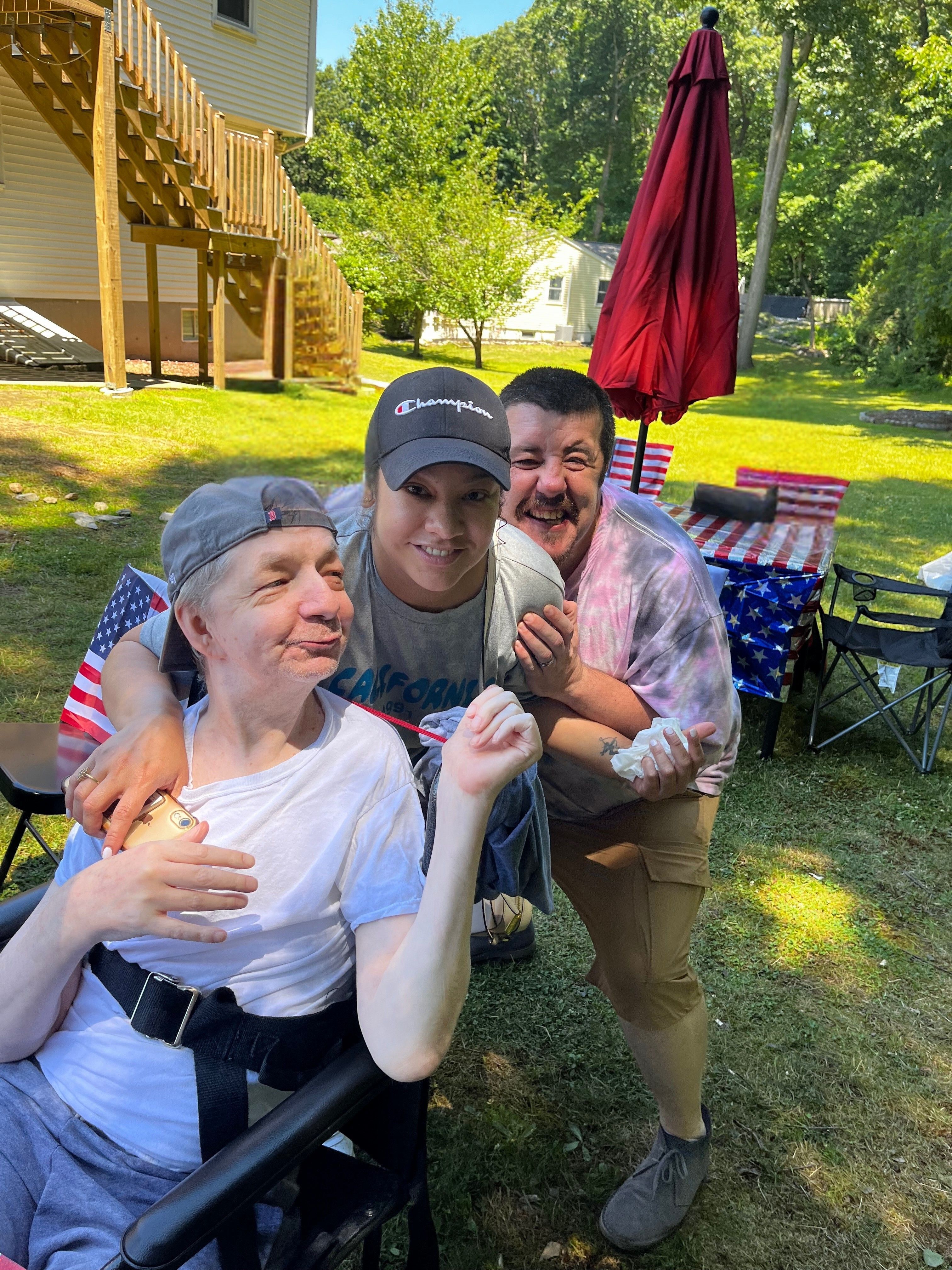 Three people smiling and embracing outdoors, with a picnic table and another person in the background.
