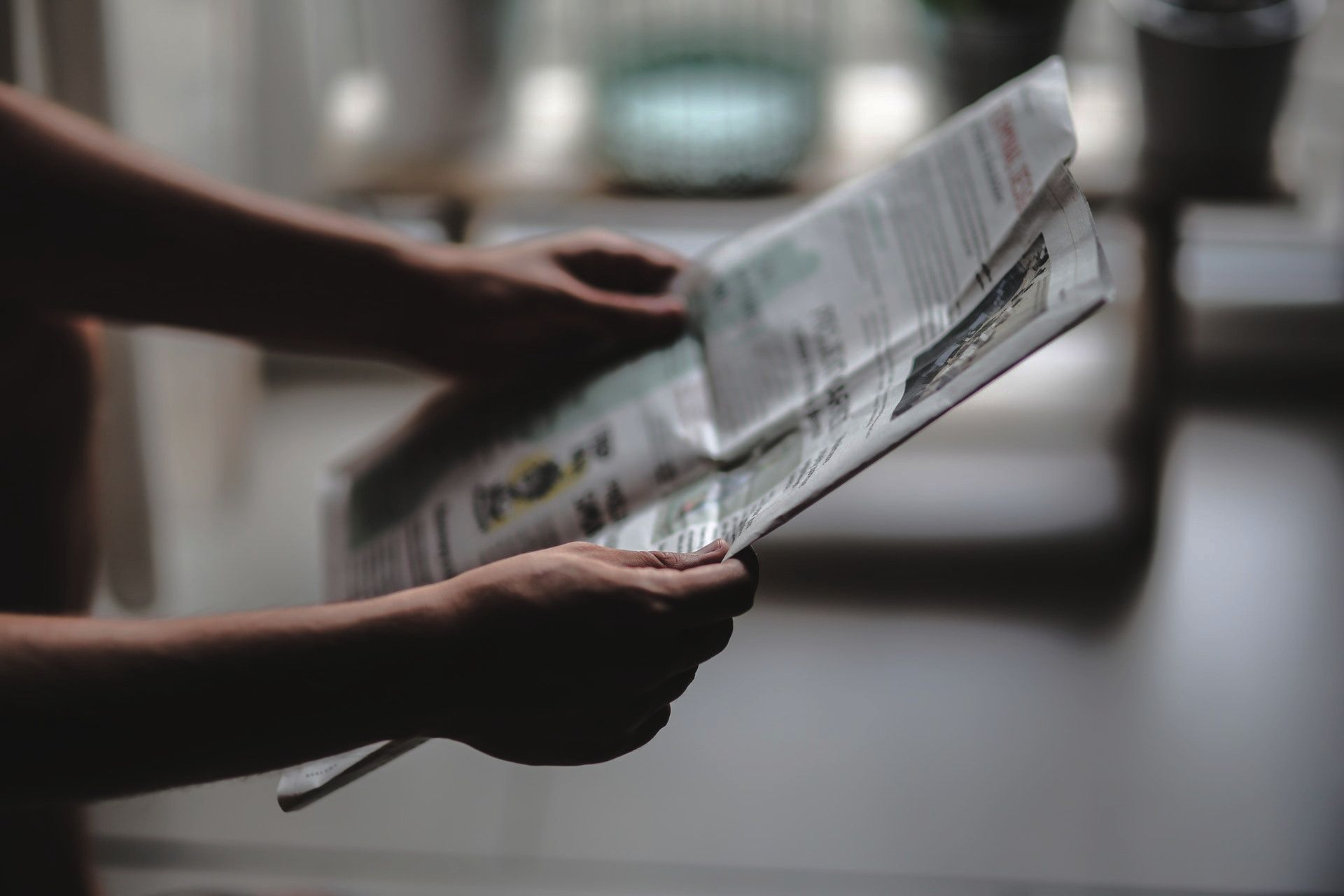 Photo of a man's forearms and hands holding a newspaper, in the foreground. The background is blurred.