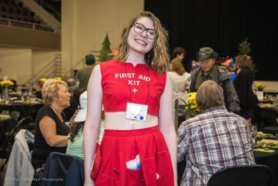 A young adult woman stands smiling at the camera. She is wearing a red costume that says First Aid. Her liver transplant scar is visible.