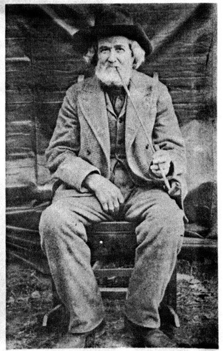 Old photo of an elderly man with a white beard, wearing a suit and hat, seated with a long pipe.