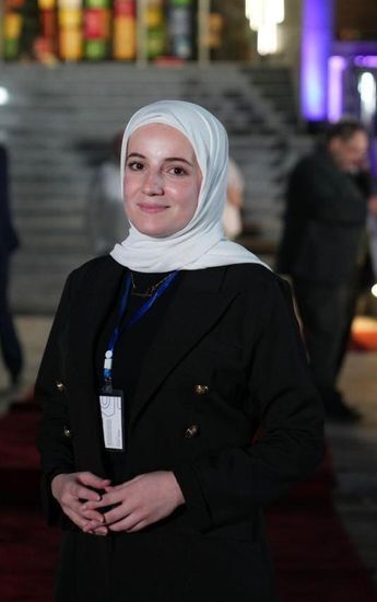A smiling woman wears a white hijab, black shirt, black blazer., with a name tag hanging around her neck. 