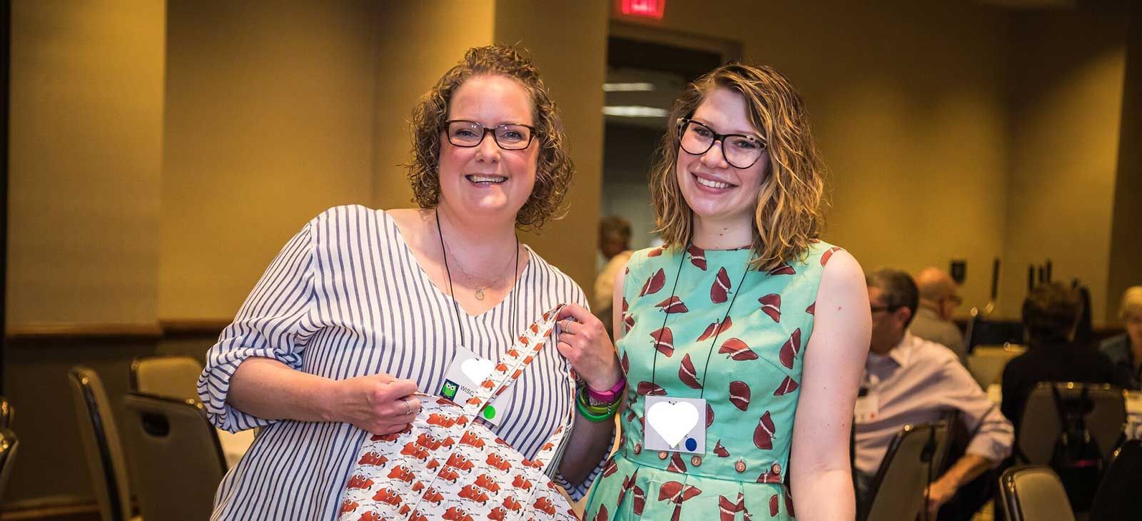 Two women look at the camera smiling. One is holding a cloth bag with cartoon livers all over it. The other is wearing a dress with cartoon livers all over it.