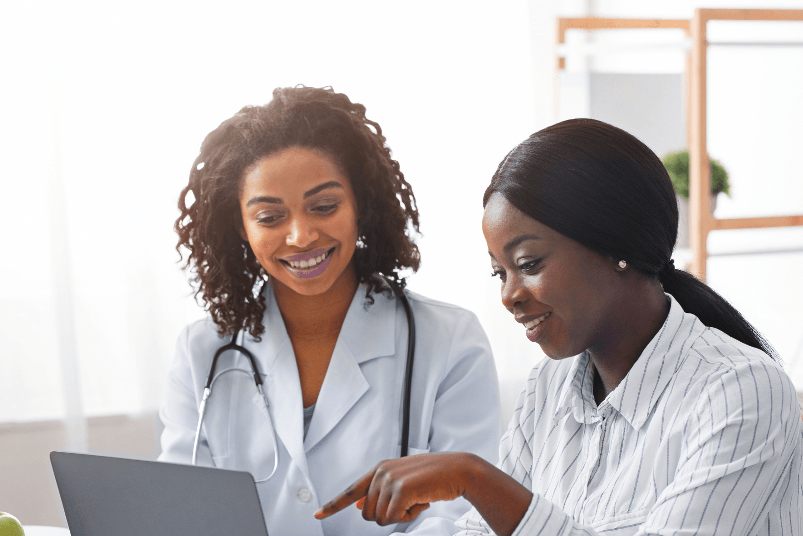 A black female doctor with curly hair compares treatment options on a computer with a black female patient with low ponytail