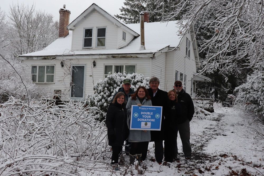 Six individuals posing outside in front of a house holding a sign that says "double your donation"