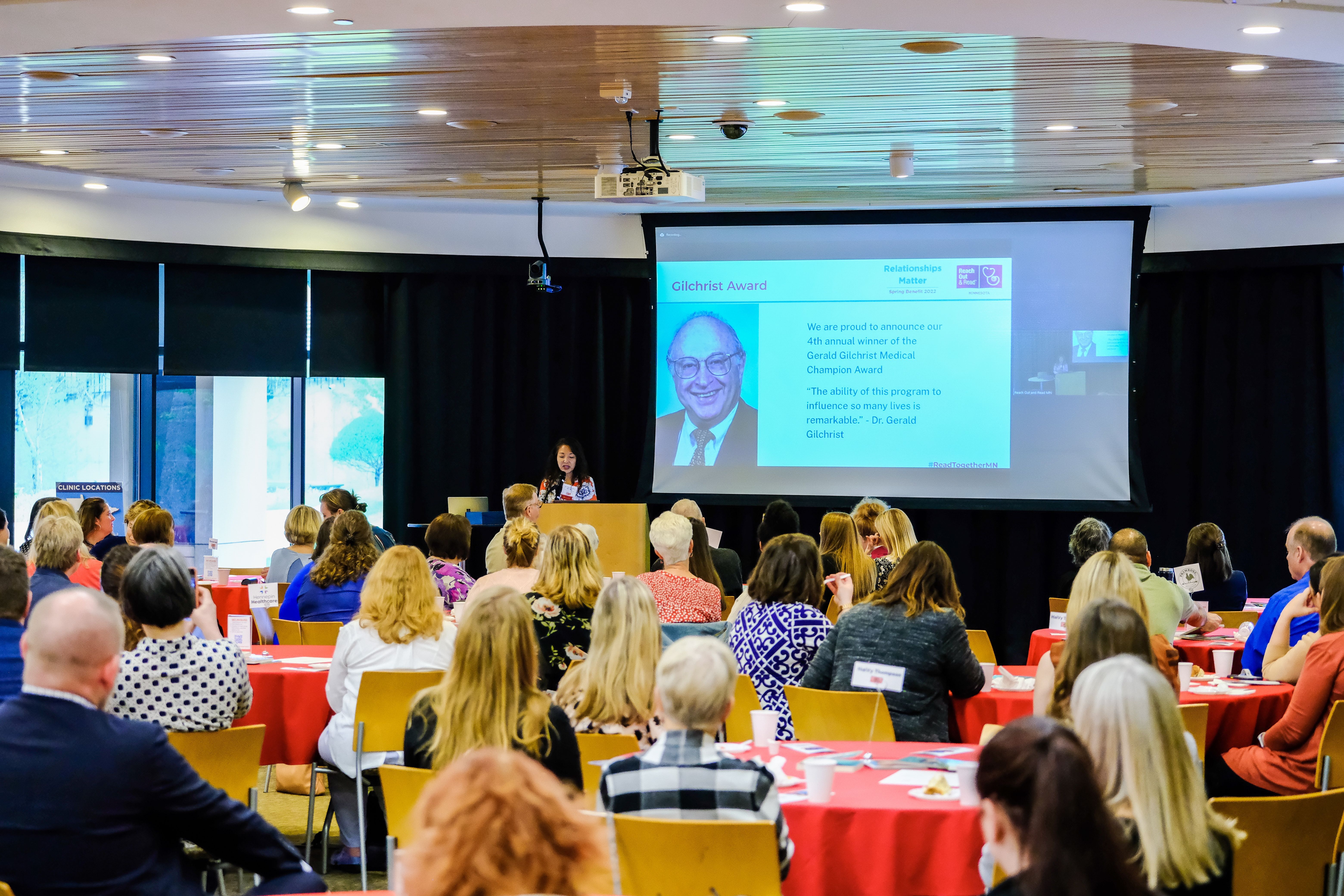 people in conference room watch woman at front podium with a projector screen