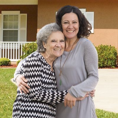 A smiling older woman hugs her caregiver in the front yard.