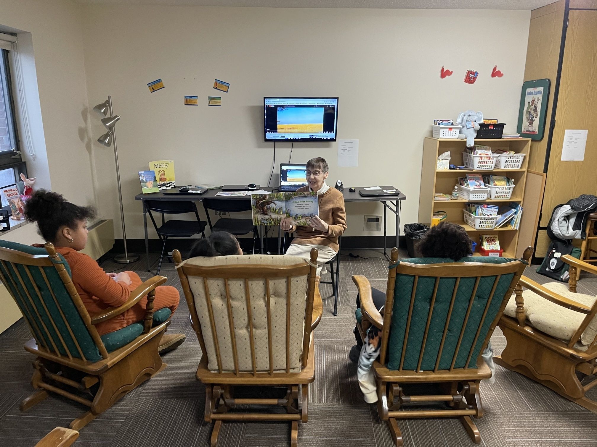 Sr. Mary Anna reads a colorful storybook to three children seated in rocking chairs. 