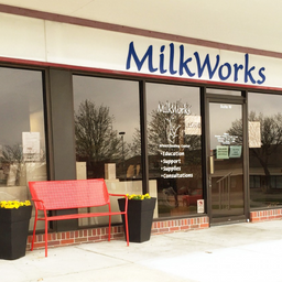 The outside of a storefront with a large blue sign that says MilkWorks. In front of the windows is a red bench with yellow flowers in black pots on either side. The curtains are open and on the window, there is white vinyl words that read "Education, Supp