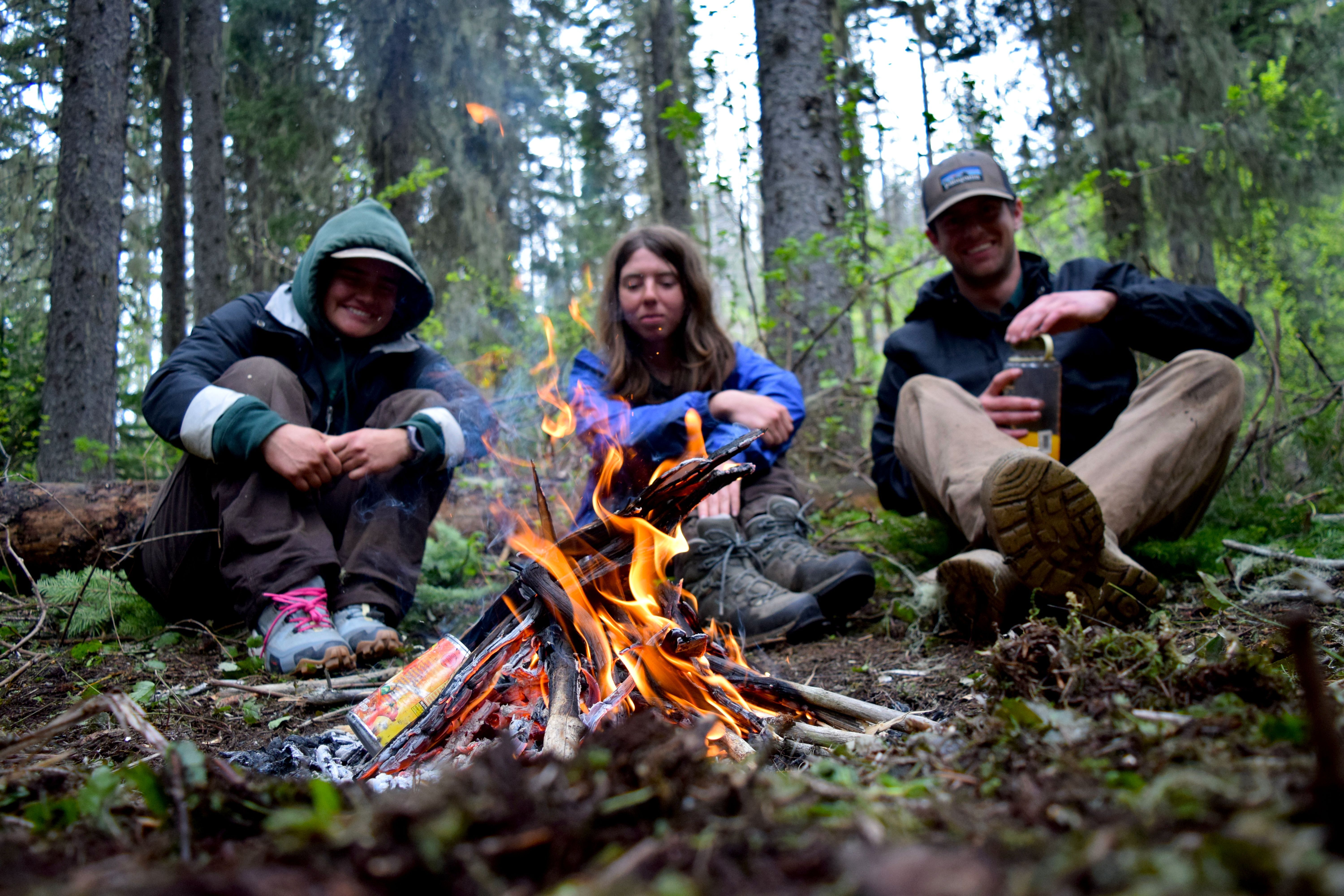 Three people are sitting on the ground behind a fire in the foreground. 