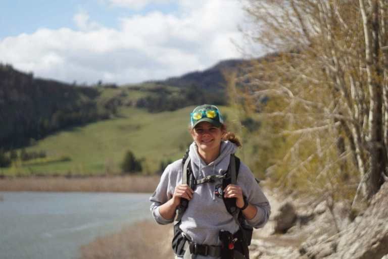 A woman stands in front of a body of water, carrying a backpack