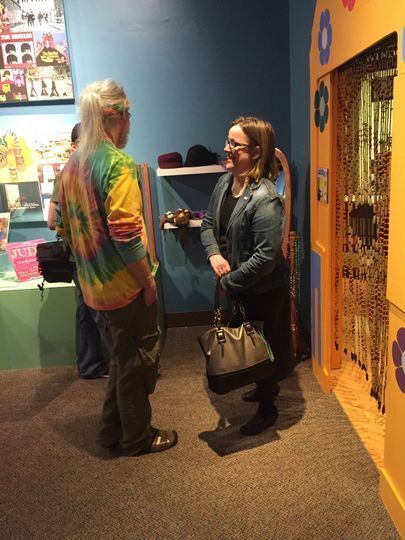 Man in tie-dyed shirt talking to a woman in a jean jacket standing in the Smoky Hill Museum's 1960s exhibit. 