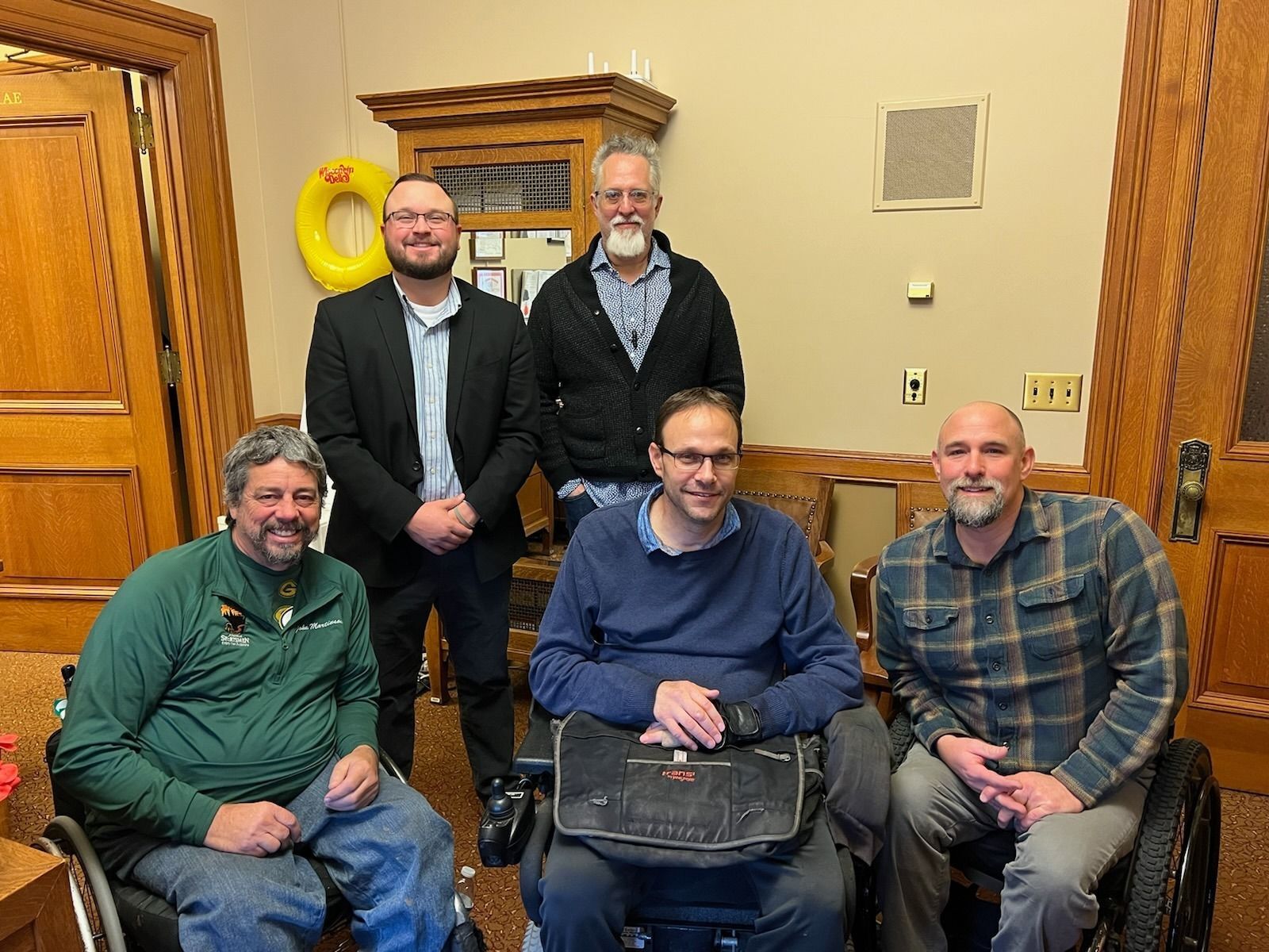 Three white men in wheelchairs sit in front of two white men standing