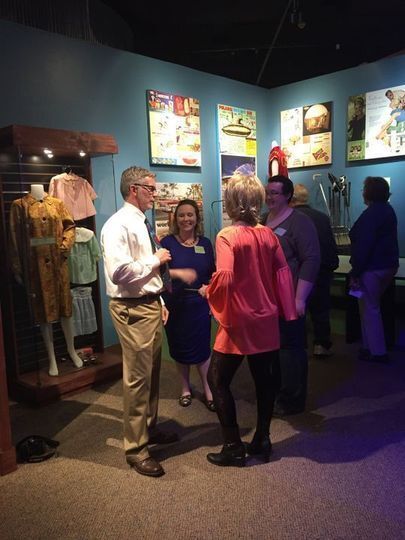 Group of people in the Smoky Hill Museum's 1960s exhibit talking. In the background is an exhibit case with 1960s women and children fashion in it.