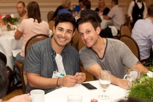 Two young adult males sit side-by-side at a conference table. They are both smiling at the camera.