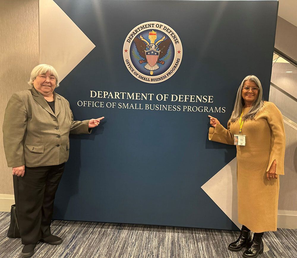 Two women stand beside a Department of Defense Office of Small Business Programs sign, smiling and pointing at it.