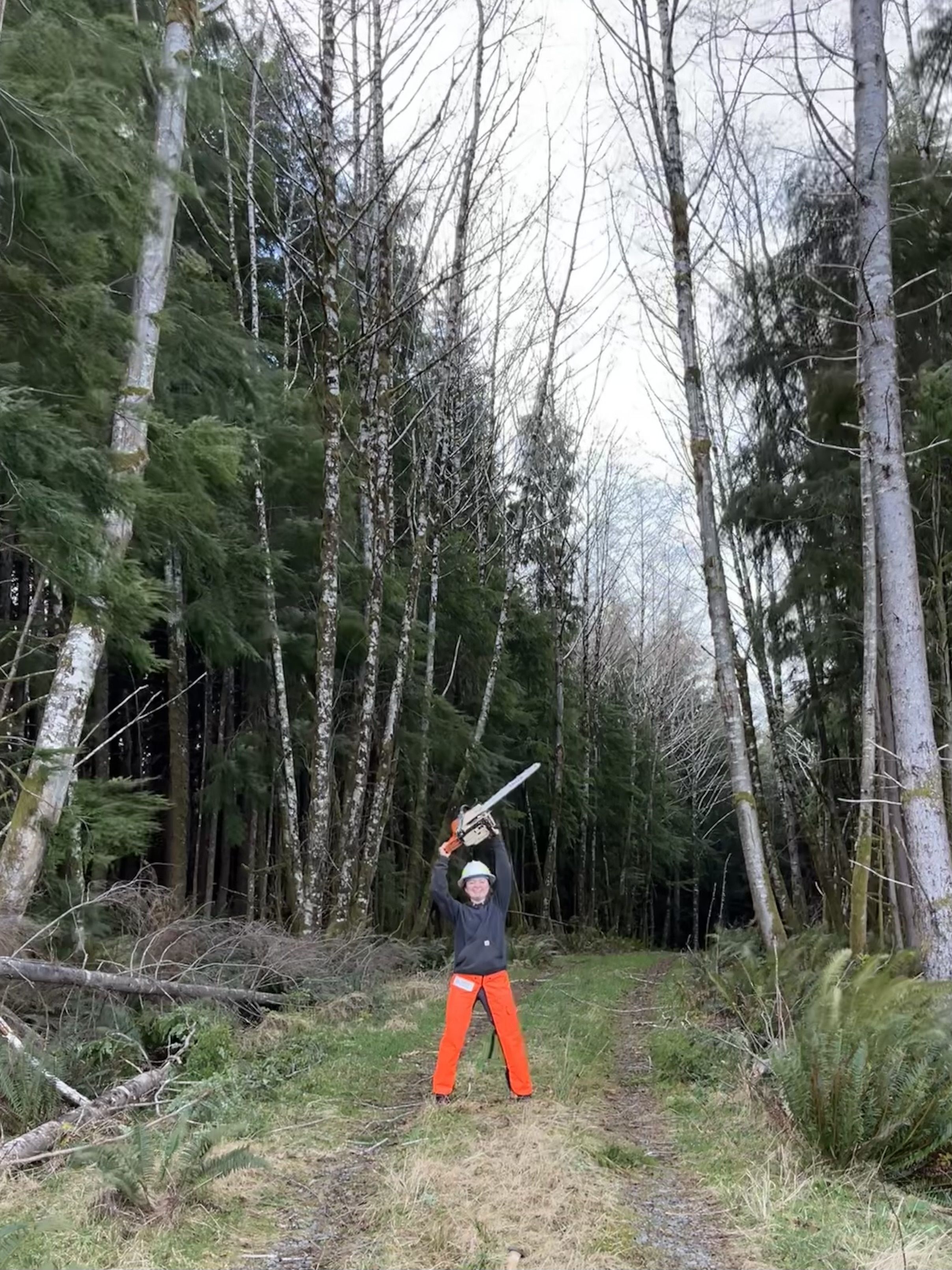 Katie stands in a forest, holding a chainsaw above her head.