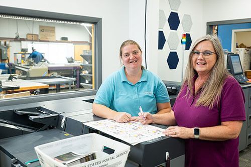 Two women standing behind a printer looking at a printed document