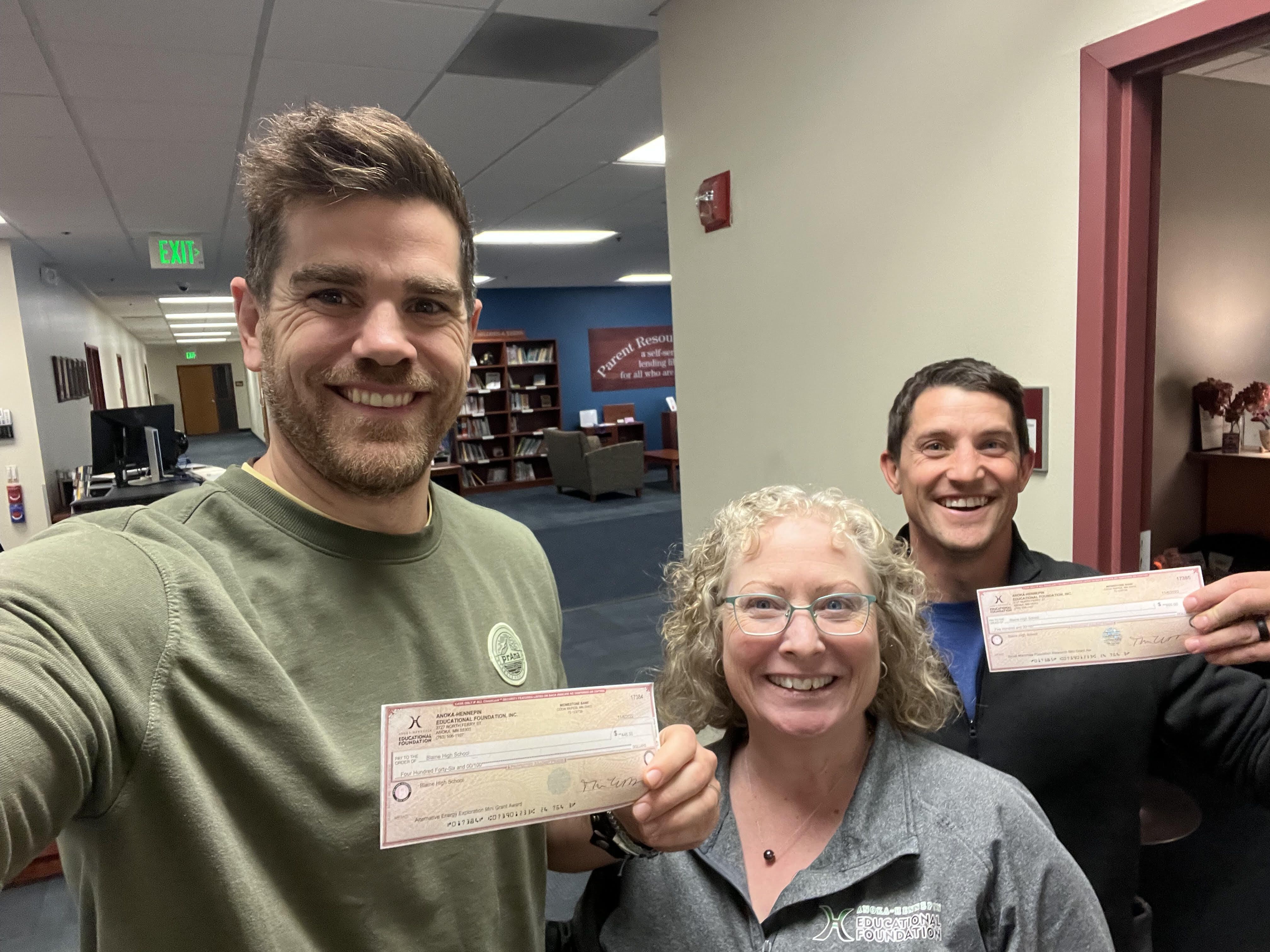 Blaine High School teachers Tim Riordan and Kendrick Novak hold up their mini-grant checks with Tess DeGeest, AHEF executive director, in the middle of the photo taken in the ESC hallway.