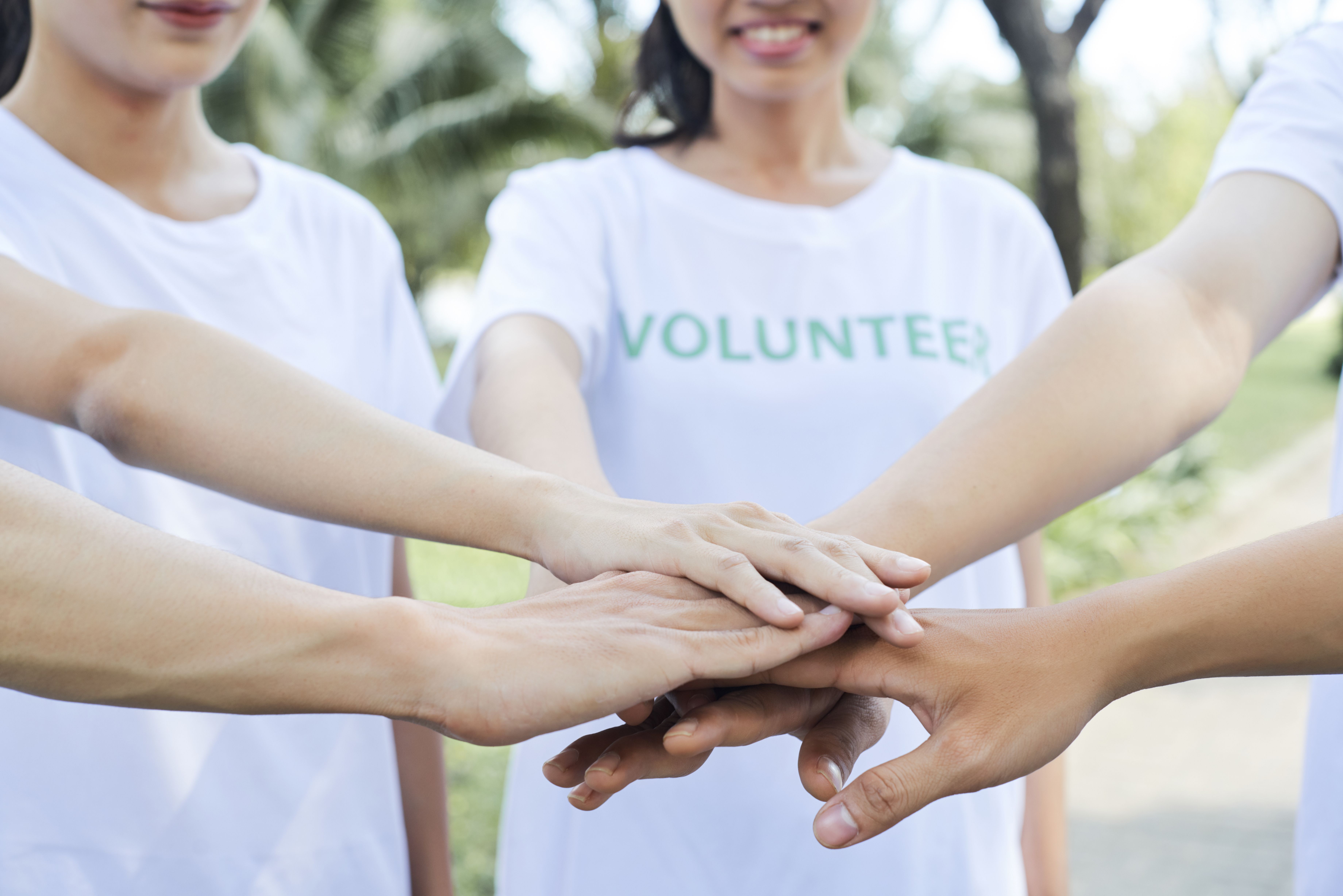 Diverse group of people holding their hands together, one wearing a VOLUNTEER t-shirt, representing teamwork and collaboration
