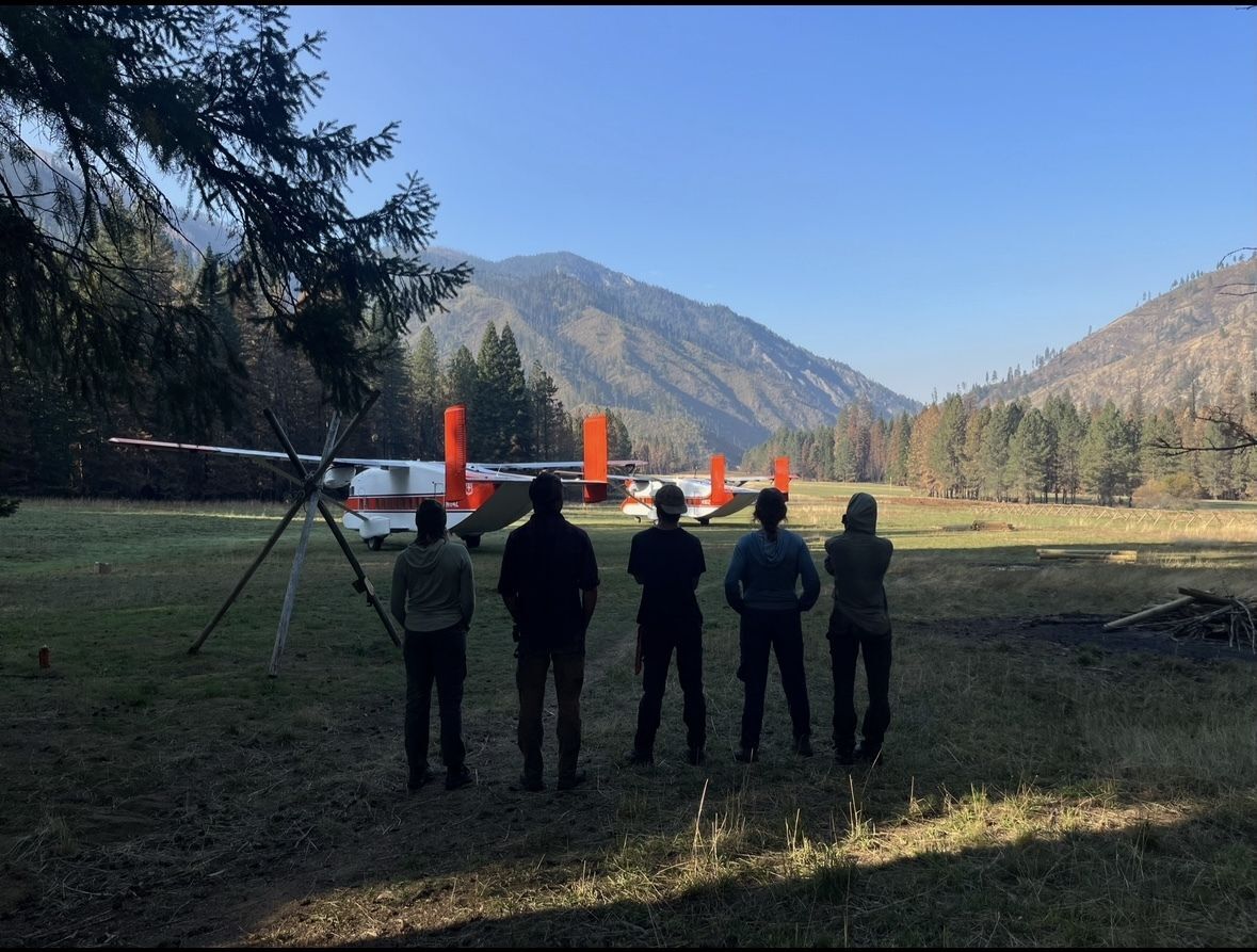 A crew stands back to the camera, with a small propeller plane in the distance.