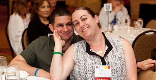 A man and woman sitting and smiling at the camera. She has her hand on his chin.