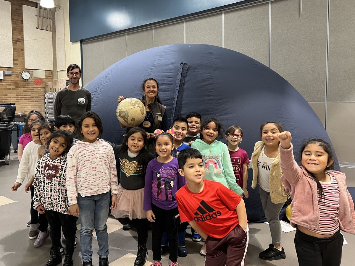 WRI Staff and a group of students excitedly pose outside the large black domed planetarium at their school.