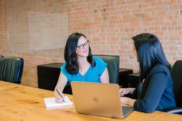 Two businesswomen sit at a table engaged in conversation with each other.  One is writing on a piece of paper and the other has a laptop in front of them.