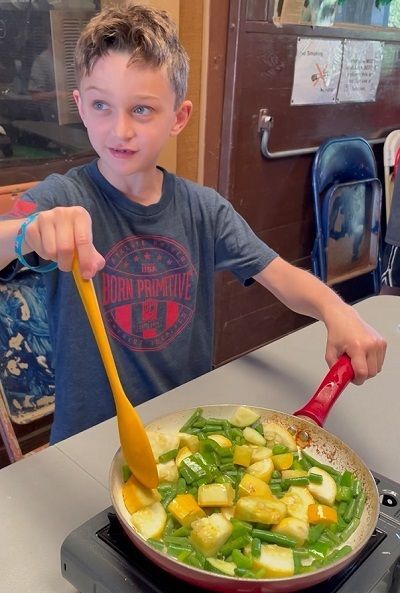 A young boy faces us holding a frying pan in his left hand and a plastic spatula in his right hand and he stirs frying vegetables