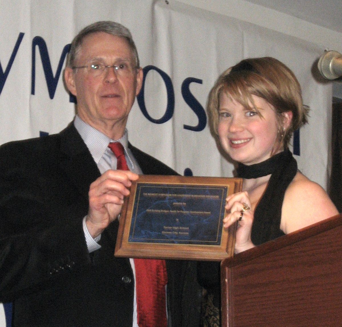 A man in a suit and red tie and a smiling woman in a black dress stand side by side on a stage, holding a wooden plaque together. Behind them, a partially visible banner hangs and a microphone stand to their right indicates a formal award presentation.