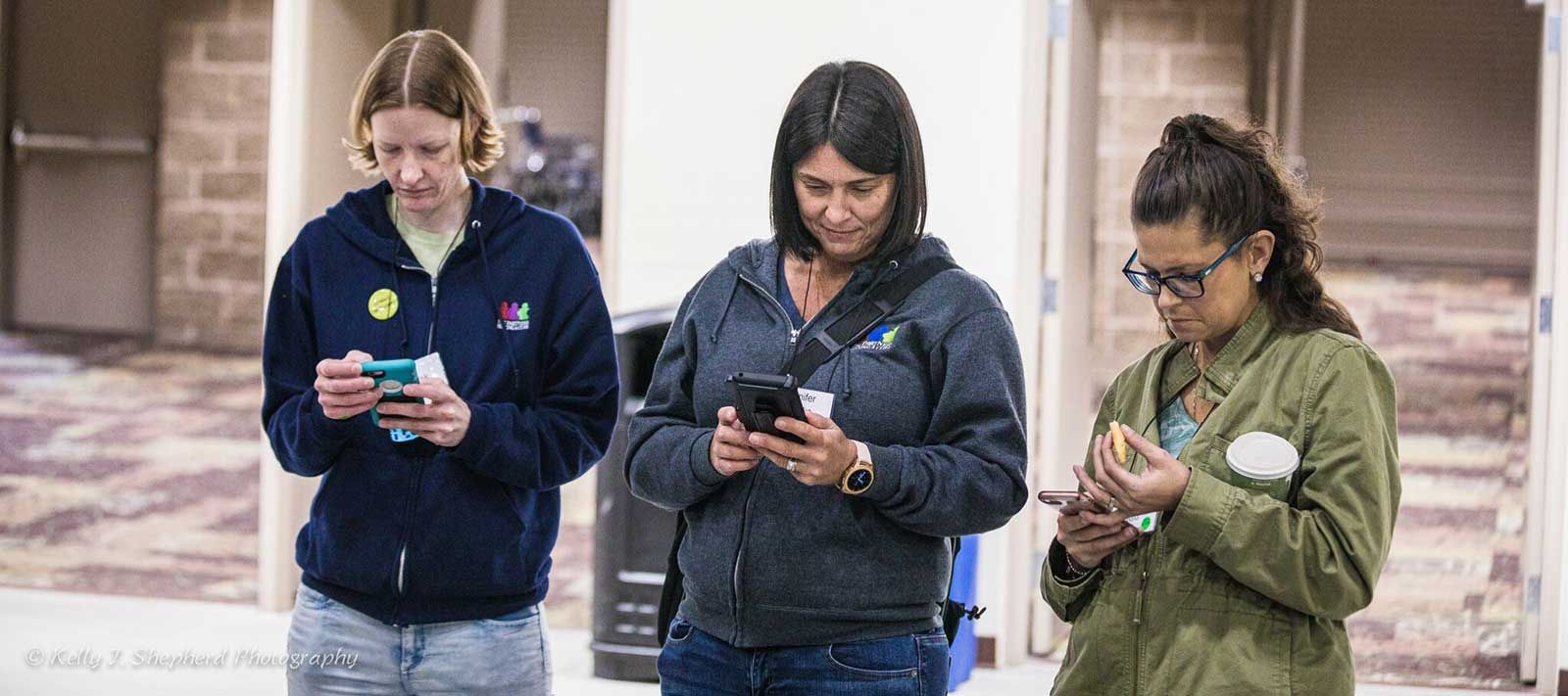 Three women stand in a row looking at their phones.