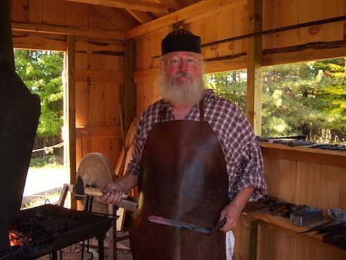 A man in a blacksmith's workshop wearing a leather apron, holding a hammer and heated metal.