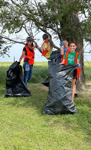 This is an image of four people standing near a tree. There is one child climbing the tree. Two children and one adult are standing on the ground with black trash bags in hand. They are all wearing safety vests. 