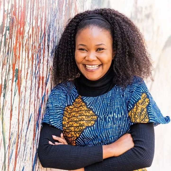 Dora Moono Nyambe smiling and leaning up against a multicolored wall.