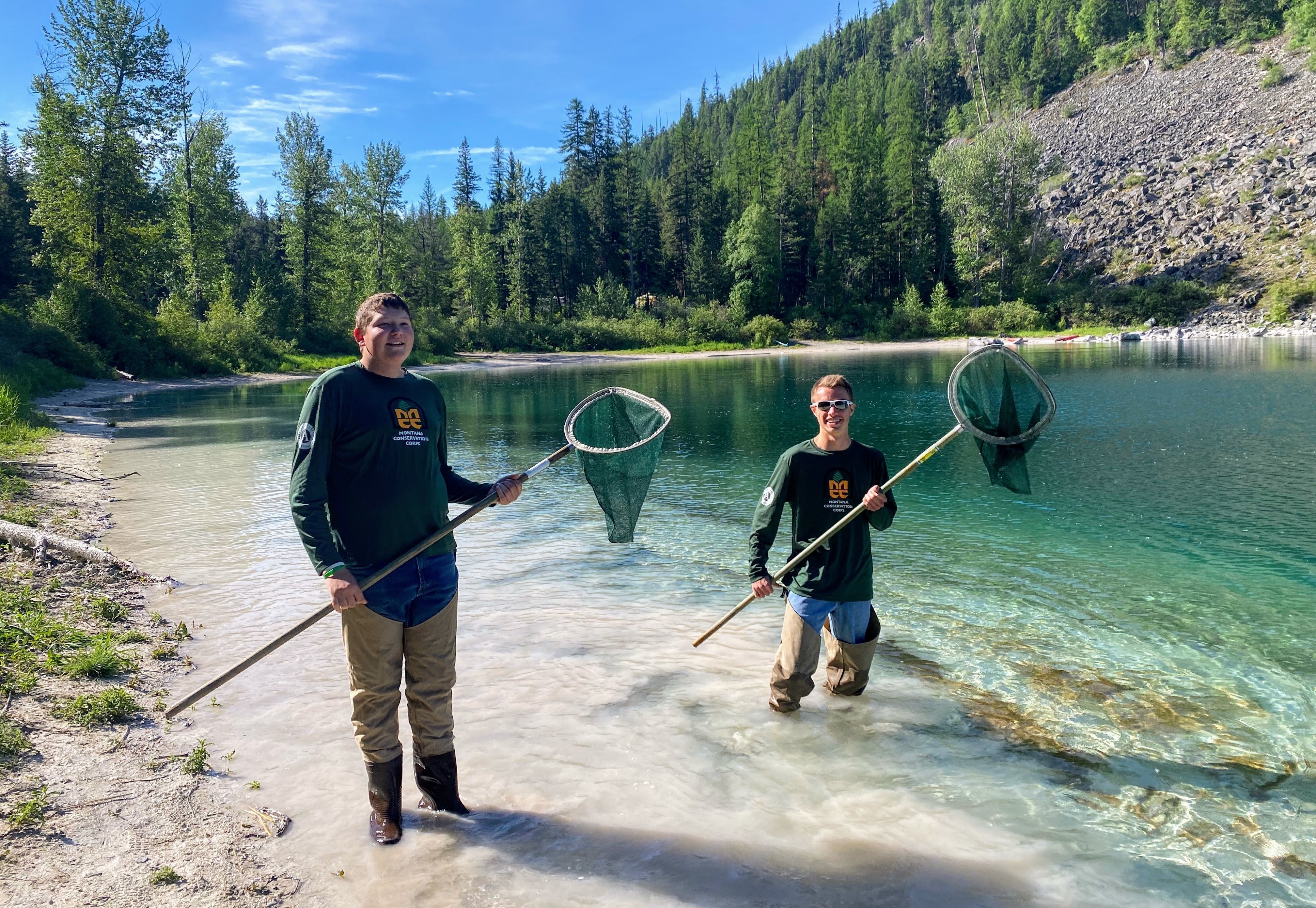 [Image Description: Two MCC members are wading into a bright blue lake. They are both wearing their MCC shirts and waders, holding nets for specimen collection.]