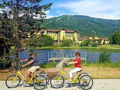Two people riding yellow tricycles on a path near a lake, with a large building and mountains in the background.
