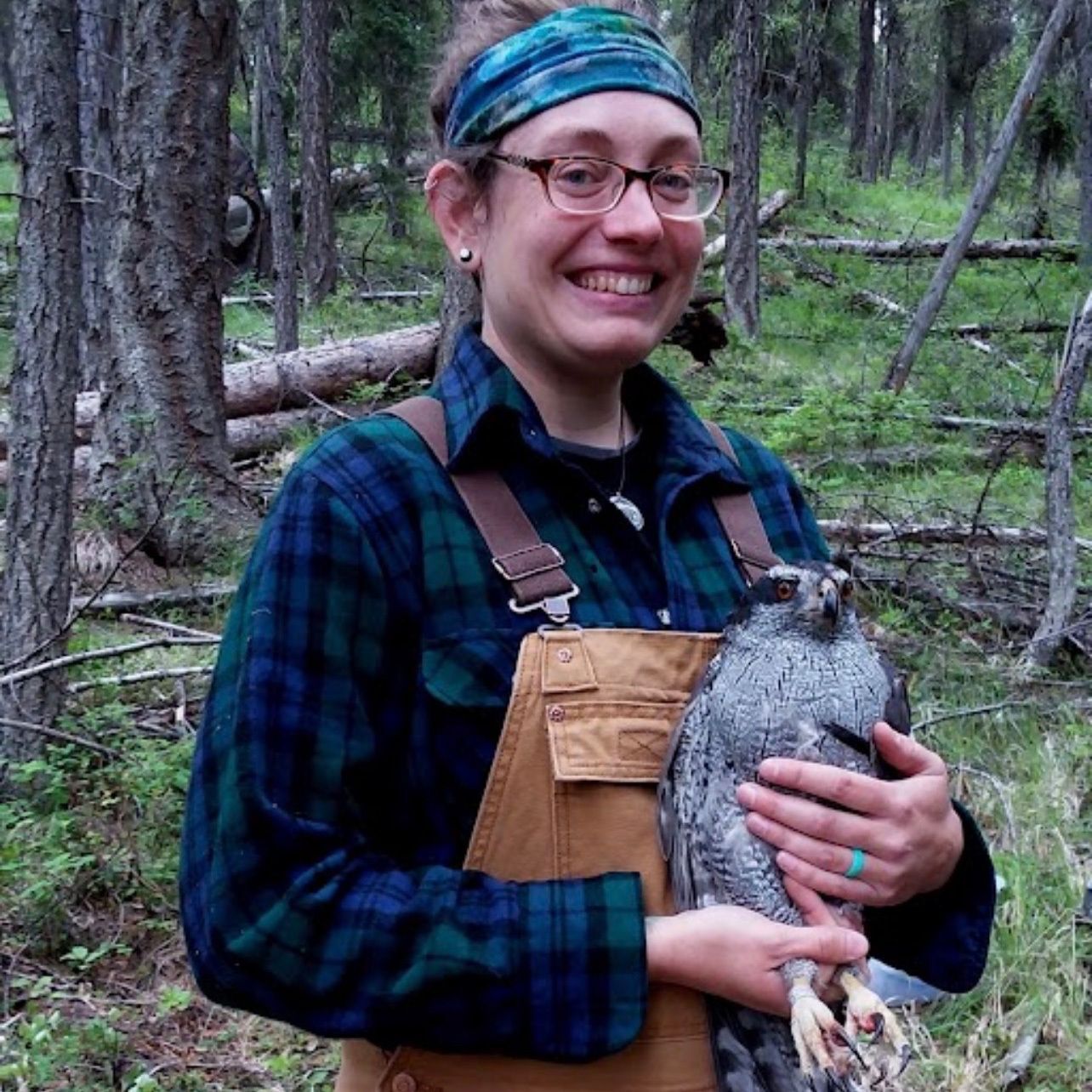 Anna Keenan holds a hawk and smiles at the camera wearing overalls and a flannel shirt