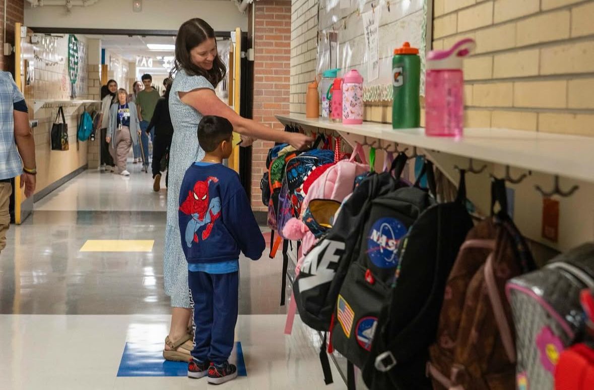 A teacher helps a young boy hang up his backpack in a school hallway on the first day of school, symbolizing care, belonging, and a fresh start.