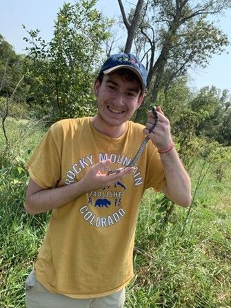 John in a yellow shirt standing in a wooded area holding a small fox snake and smiling