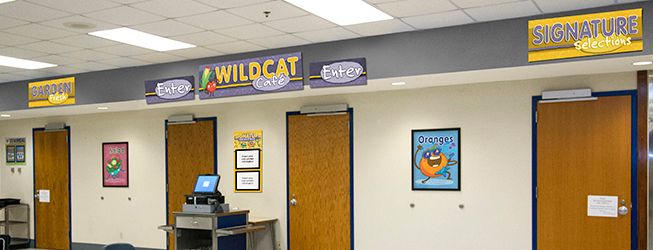 Cafeteria signs above entrance doors, purple and gold with food characters, menu board, line signs