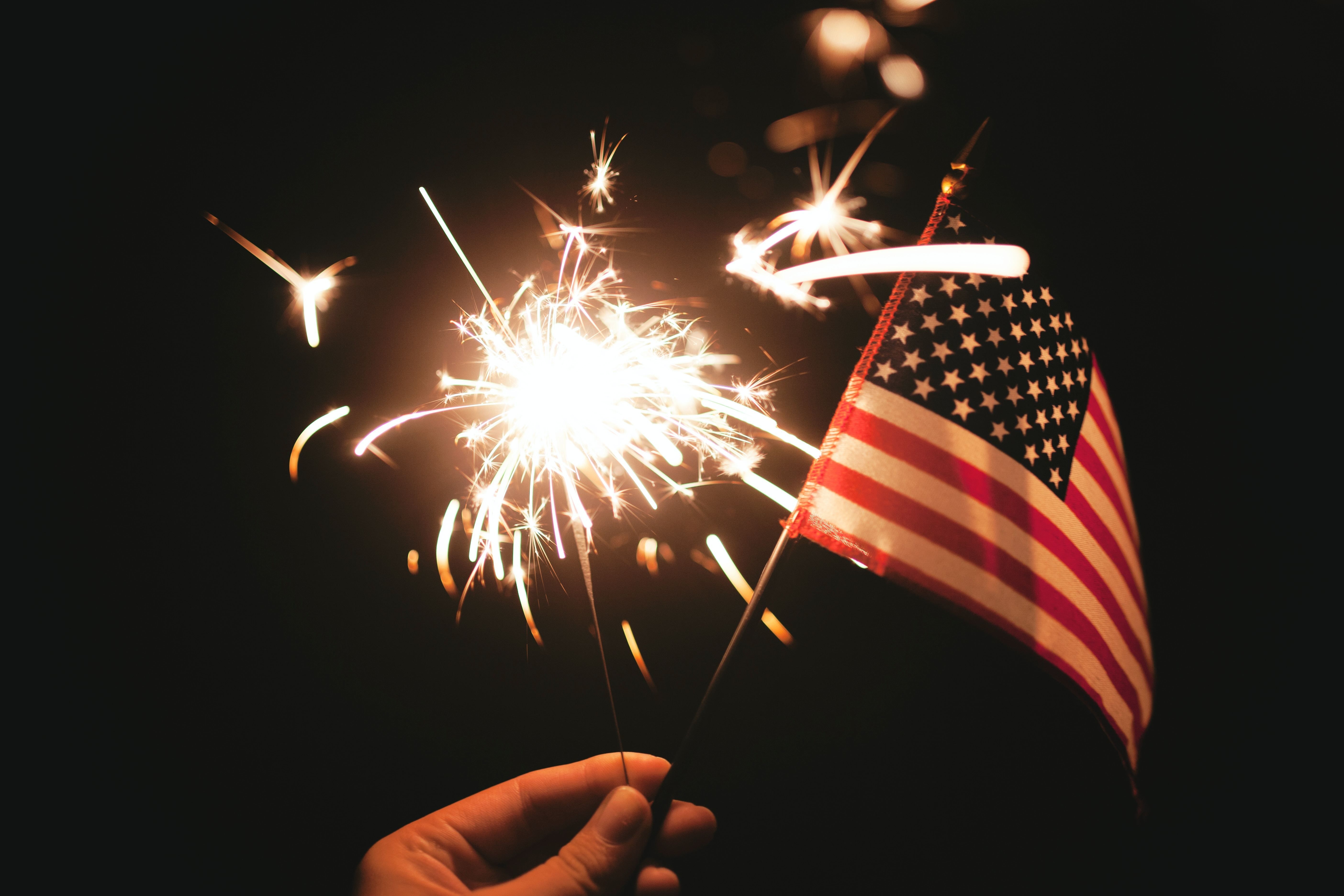 A night sky lit up with fireworks. There is a hand waving a small, American flag.